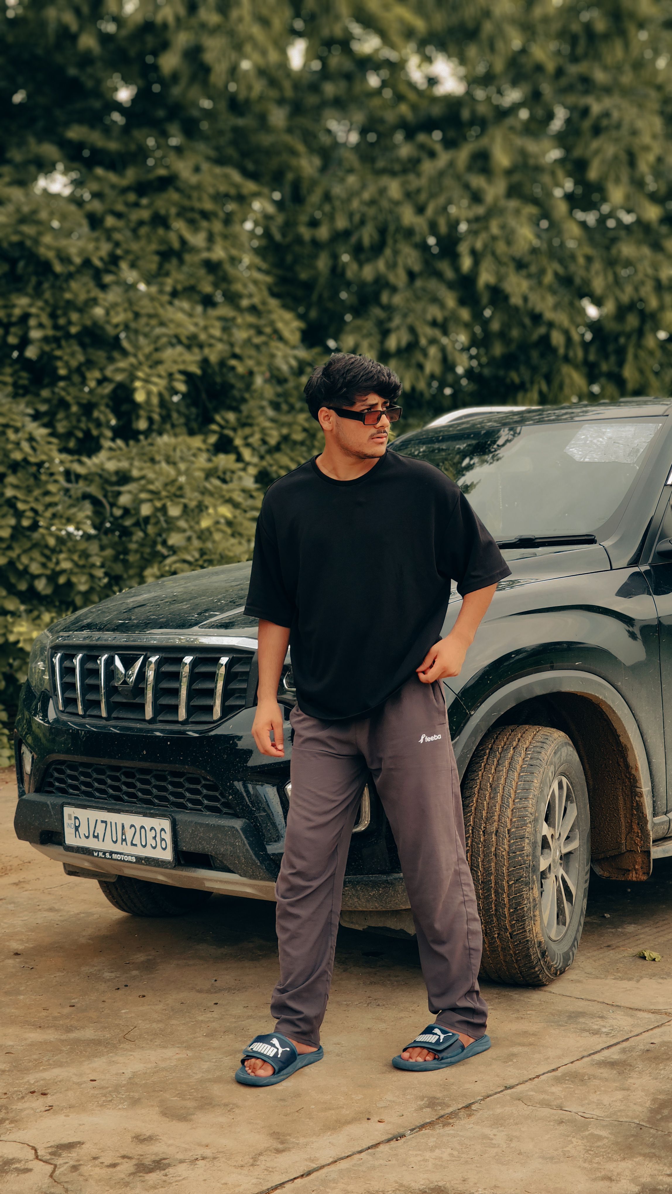 A man in casual wear stands beside a black SUV outdoors, with greenery in the background.