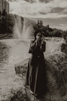 Elegant black and white portrait of a woman holding a camera by a scenic fountain outdoors.