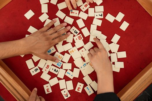 Three hands engage in a tile-based board game on a vibrant red surface.