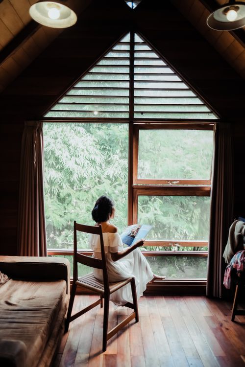Free Woman reading by a large window in a cozy wooden cabin, enjoying a peaceful moment. Stock Photo
