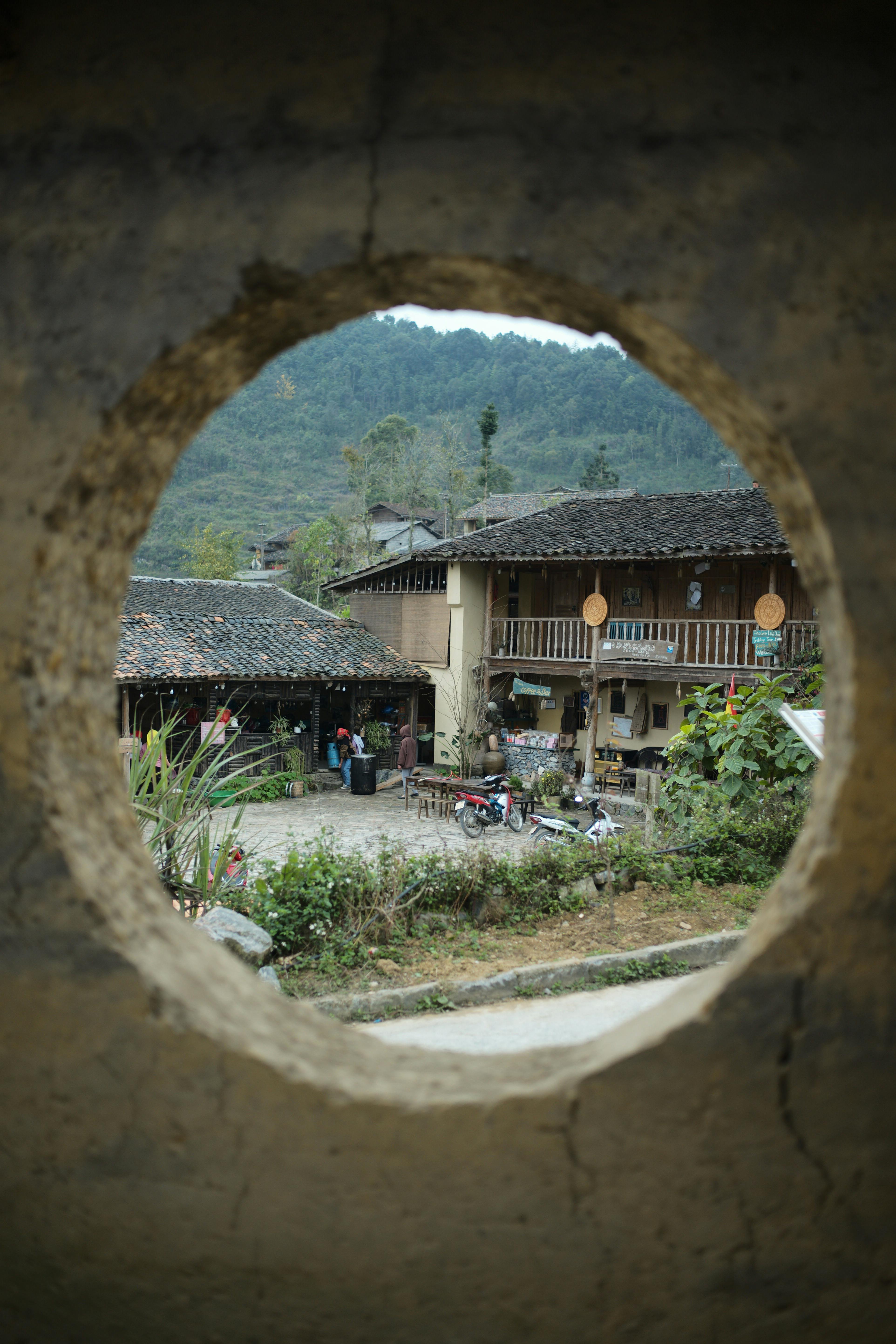 Charming view of a traditional building in Ha Giang, framed through a round window with scenic mountains.