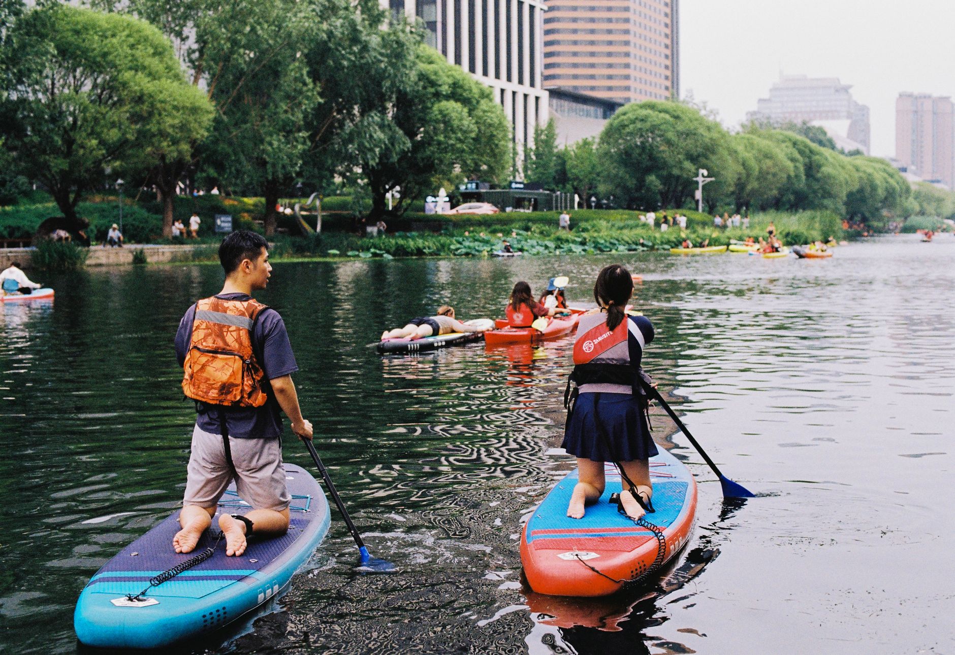 A group of adults and children paddleboarding on a calm river in a city park.