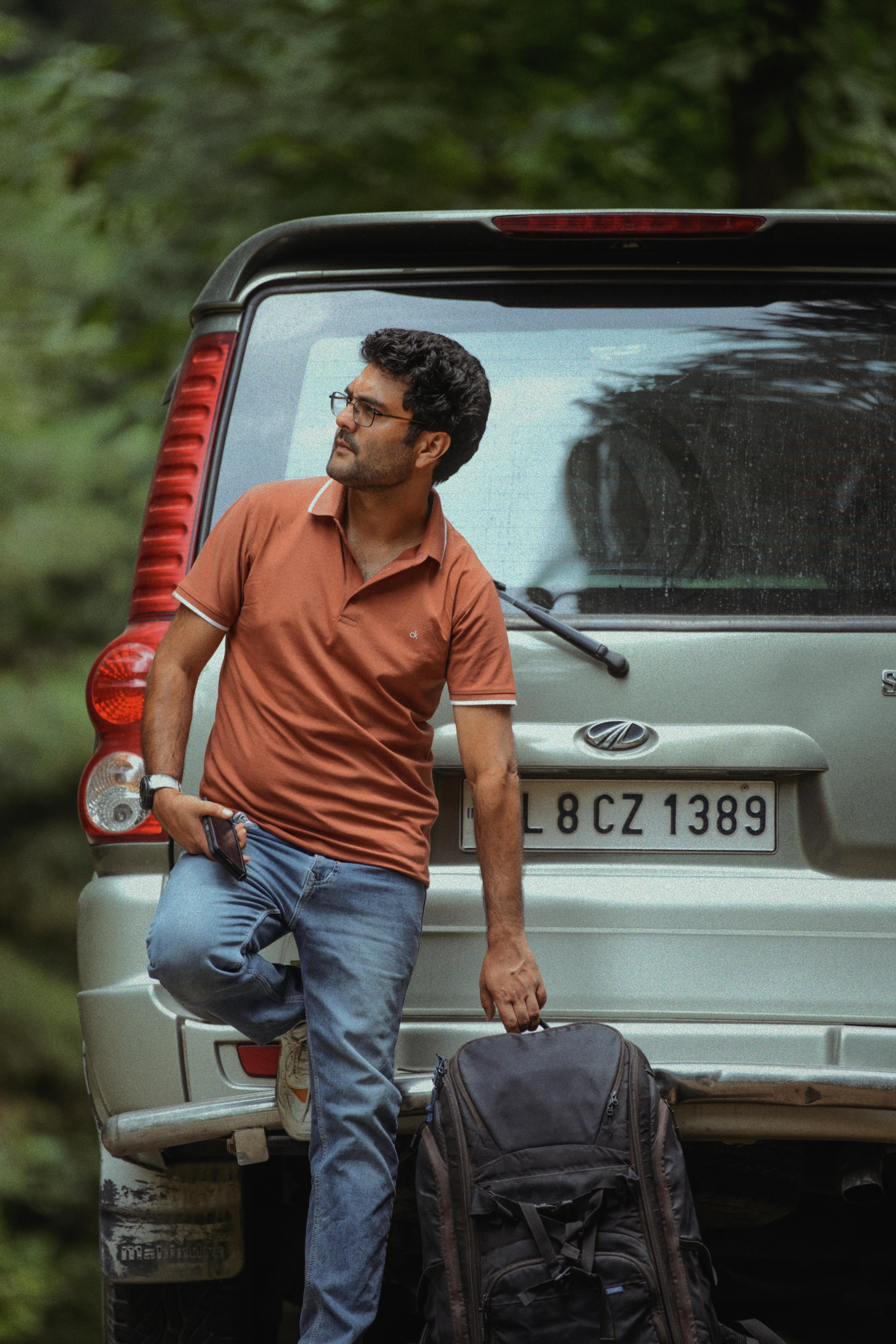 A man in casual attire leans against an SUV parked in a forest setting, embodying travel and adventure.