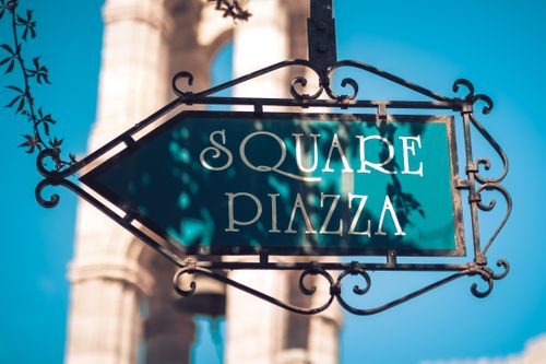 Free Elegant vintage square piazza sign with ornate iron design, set outdoors under bright sunlight. Stock Photo