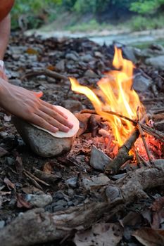 A person cooks a tortilla on a rock by a campfire in a forested area, evoking a sense of rustic outdoor adventure.
