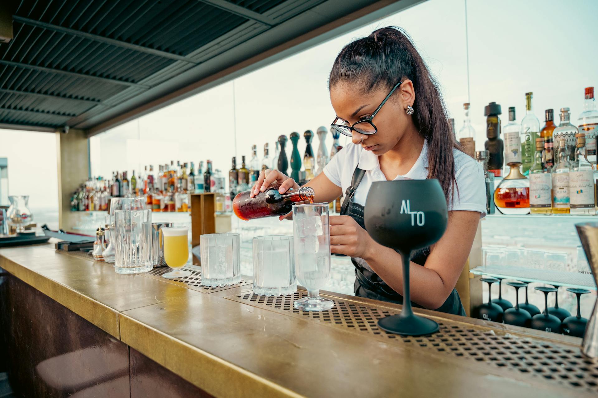 A female bartender skillfully prepares cocktails at a stylish rooftop bar with skyline views.