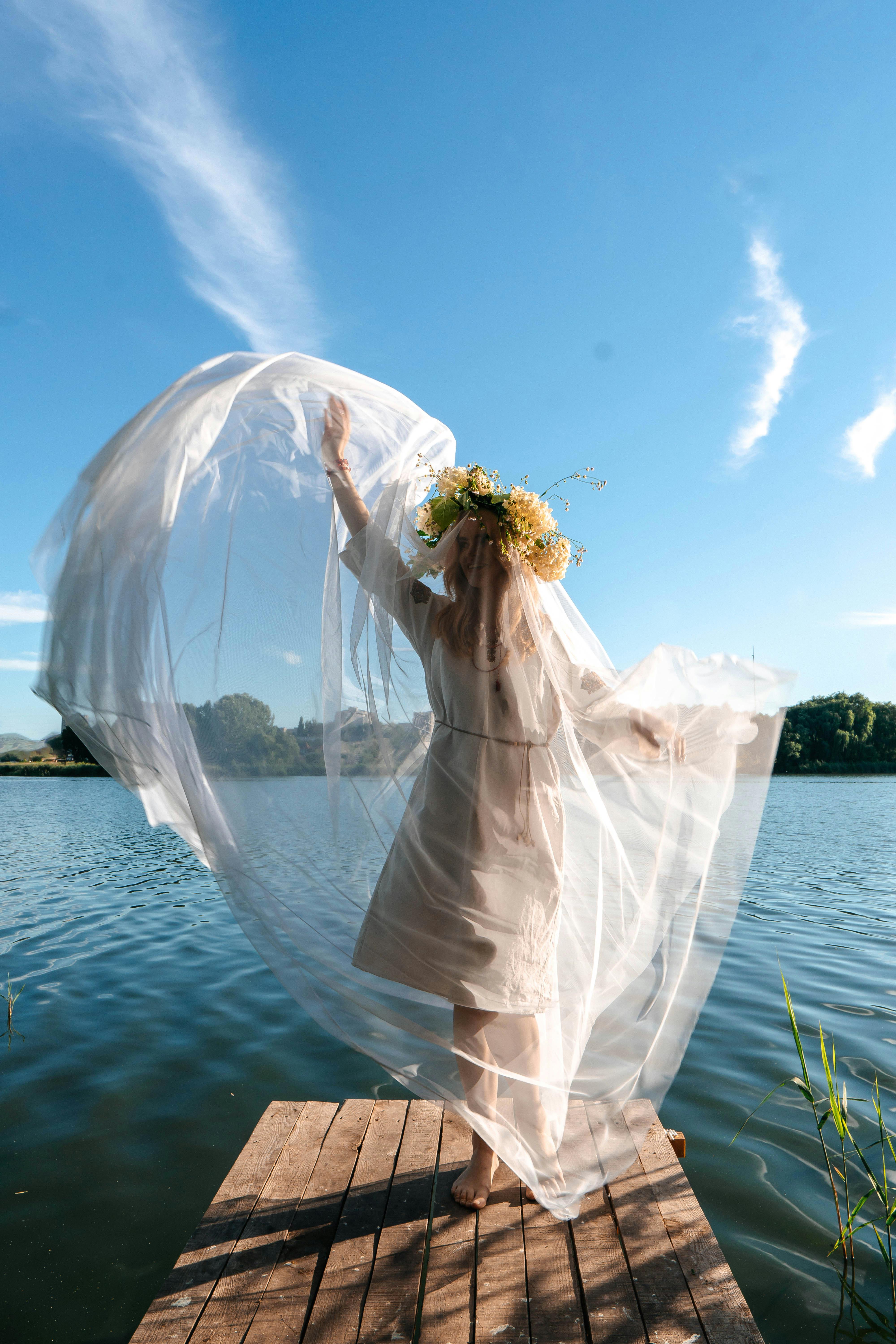 Caucasian woman in white dress with floral headdress enjoys nature on a sunny day by the lake.