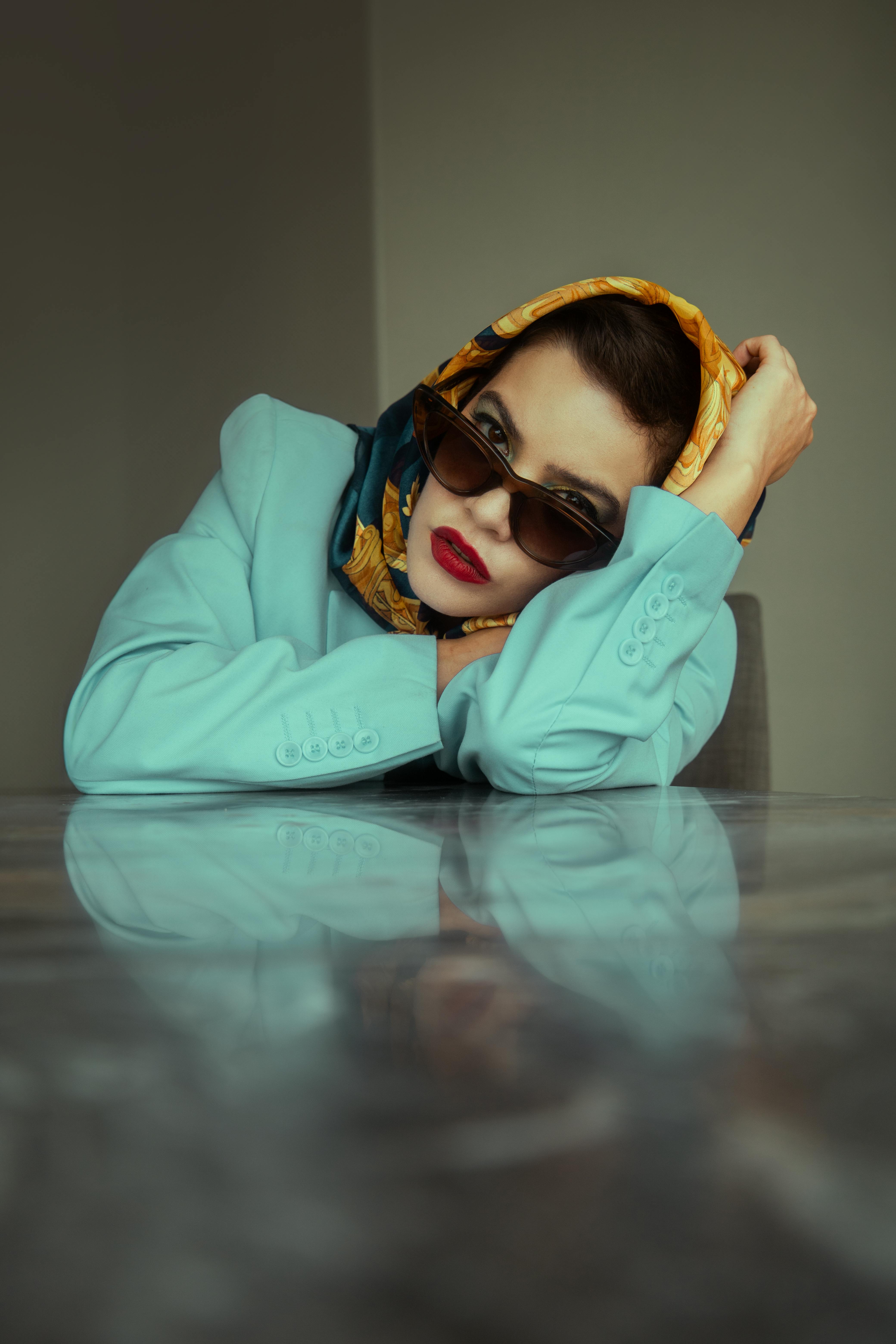 Free Chic woman in sunglasses and headscarf poses indoors with a reflective table. Stock Photo