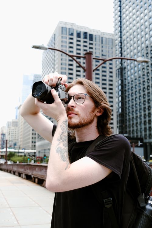 Free A young male photographer with eyeglasses captures cityscape using a vintage camera outdoors. Stock Photo