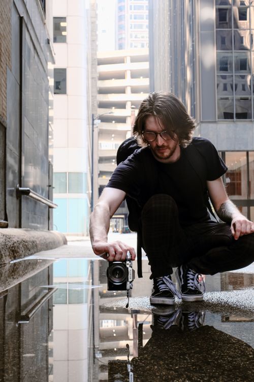 Free A young man takes a photo in a city setting, focusing on reflections in a puddle. Stock Photo