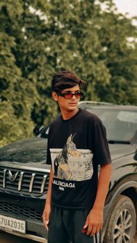 Teenager posing with car wearing sunglasses and fashion t-shirt, captured outdoors in summer.