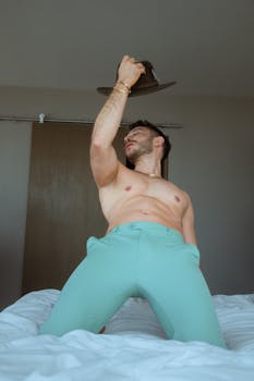 Shirtless man in stylish pants poses in a modern bedroom, showcasing a blend of elegance and casual fashion.
