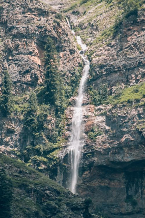 Free A breathtaking waterfall cascades down a rocky cliff, surrounded by lush greenery. Stock Photo