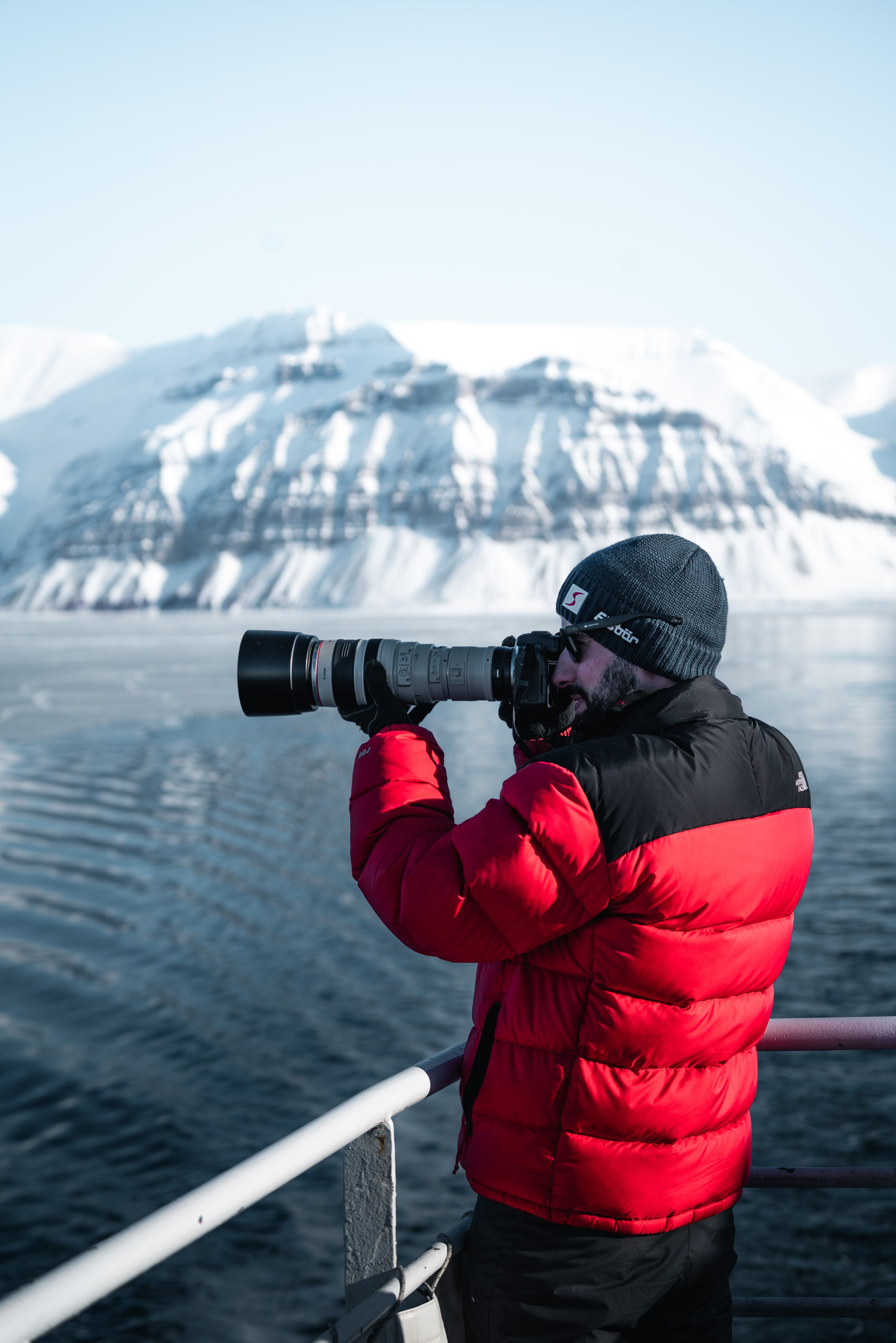 A man in a red jacket photographs snowy mountains on an Arctic expedition.