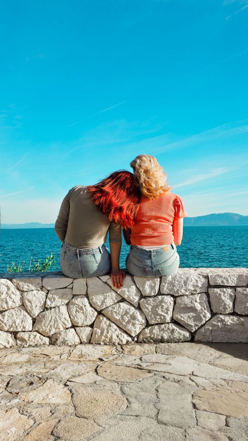 Free Two women sit together on a stone wall by the ocean, enjoying a sunny day. Stock Photo