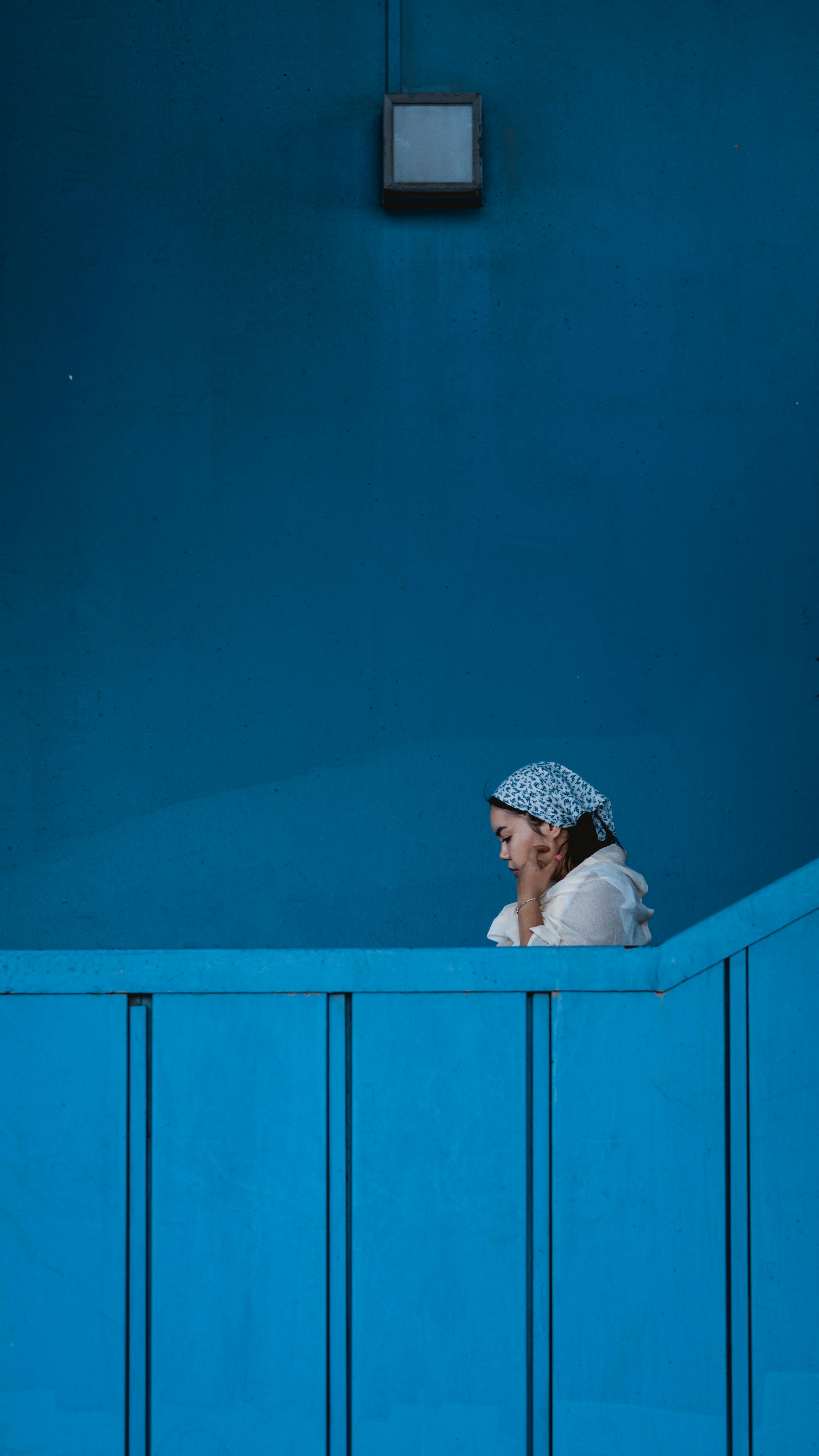 A woman in a blue headscarf is leaning against a vibrant blue wall indoors.