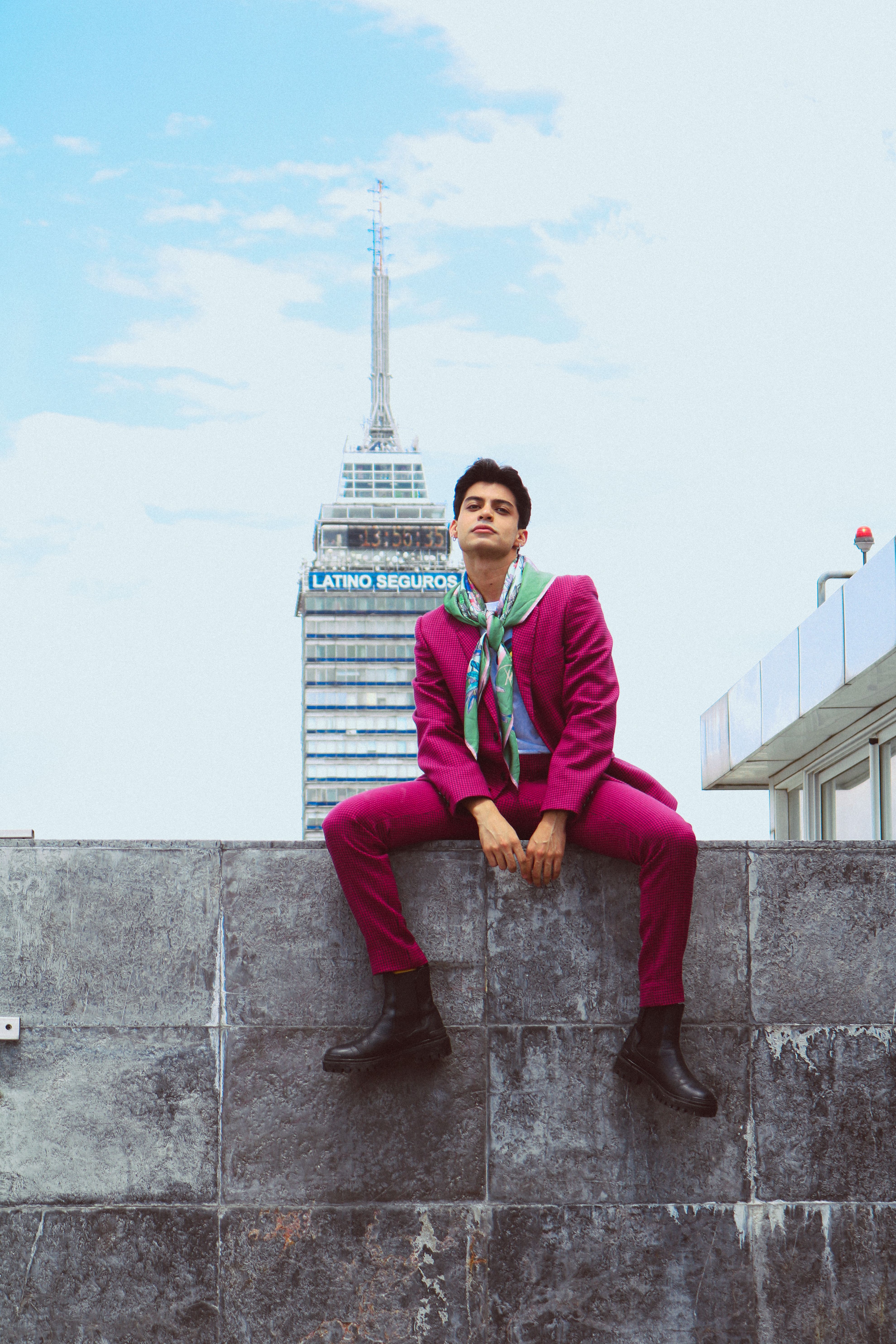 Young man in vibrant suit seated outdoors with Torre Latinoamericana in background.