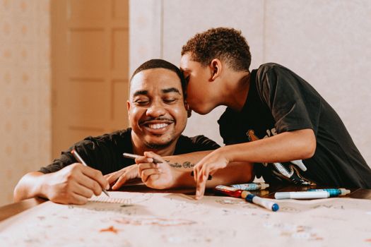 A joyful father receives a kiss on the cheek from his son as they engage in drawing together indoors.