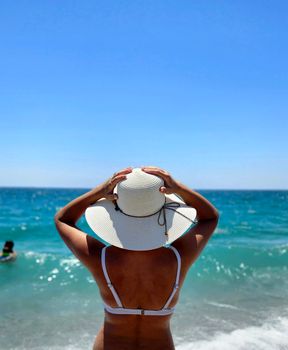 A woman in a bikini and sun hat enjoying a sunny day at the beach with ocean waves.
