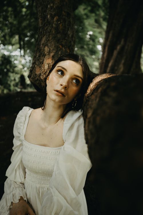 Free A contemplative young woman in a white dress leaning on a tree in a lush forest, with a soft, dreamy expression. Stock Photo