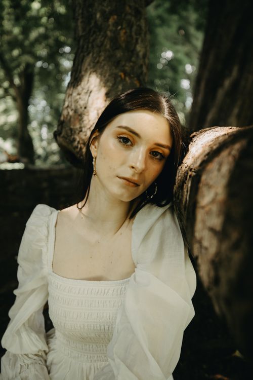 Free A serene portrait of a woman in a white dress leaning against a tree in a sunlit forest. Stock Photo