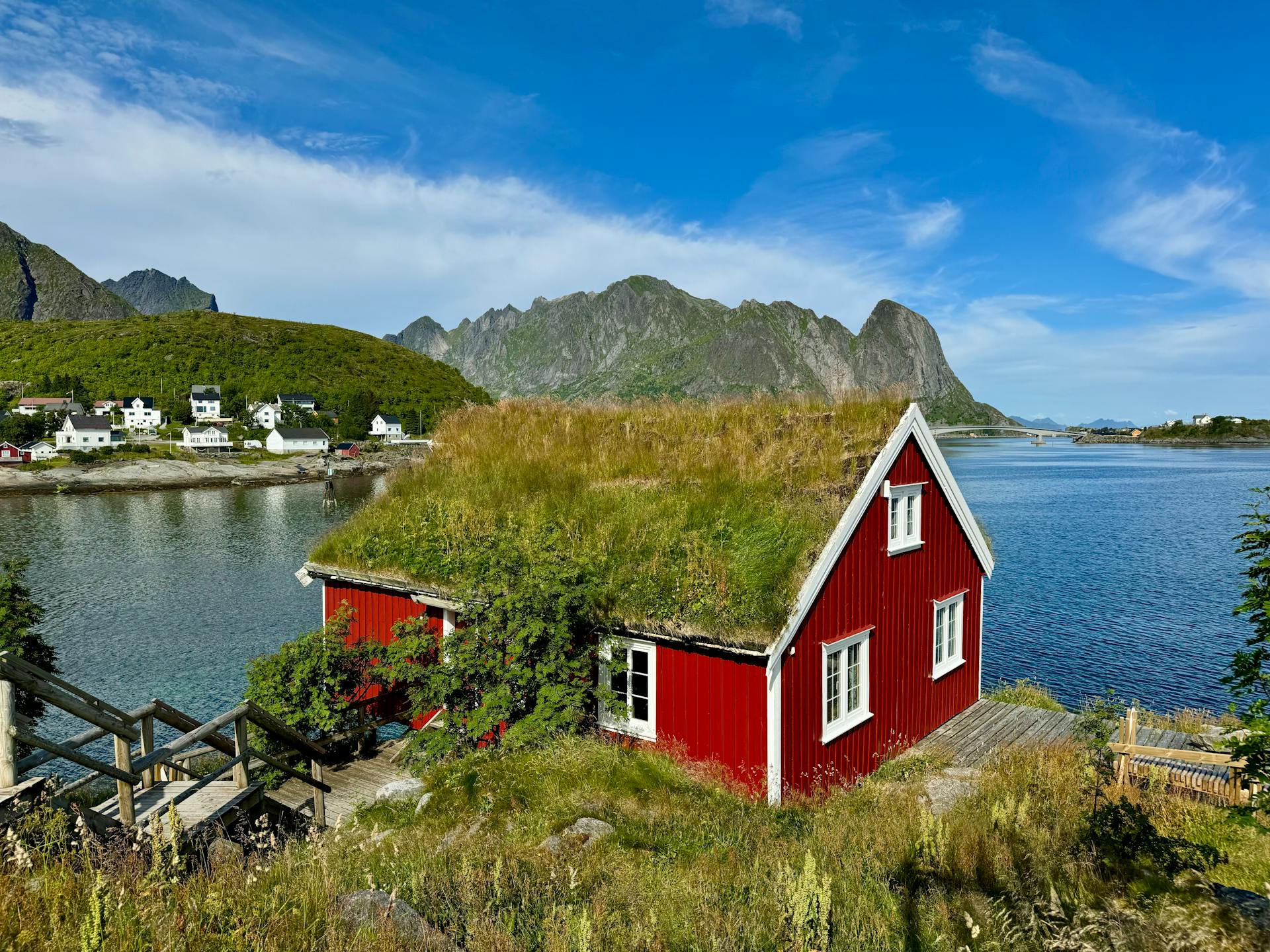 Charming red cabin with grass roof by a lake, surrounded by Nordic mountains and nature.