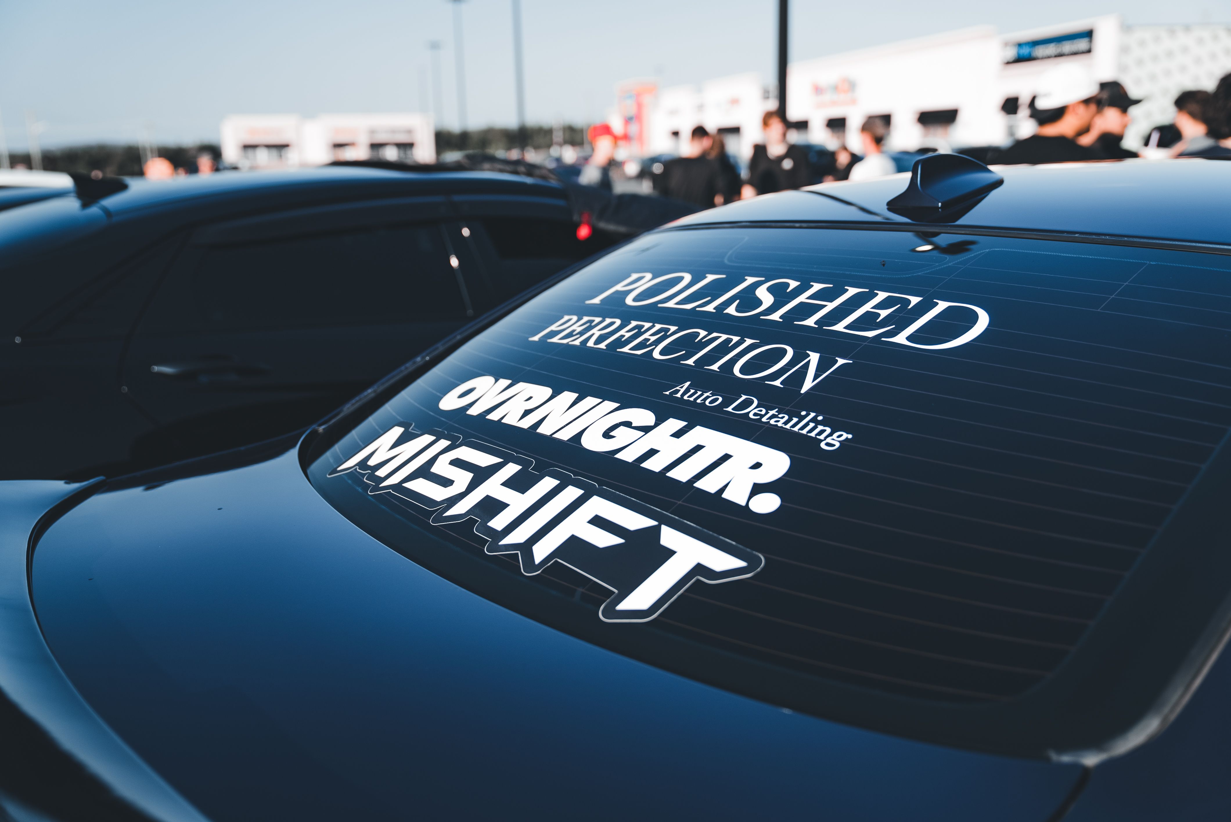 Rear view of black car with custom window decals at an outdoor auto event.