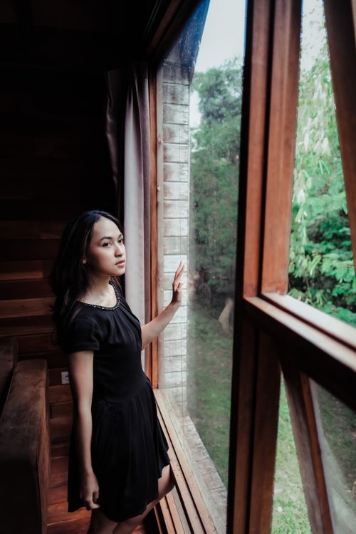 Free A young woman in a black dress looks thoughtfully outside through a large wooden window. Stock Photo