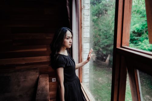 Free A thoughtful woman in a black dress gazes out a wooden window, exuding elegance and mystery. Stock Photo