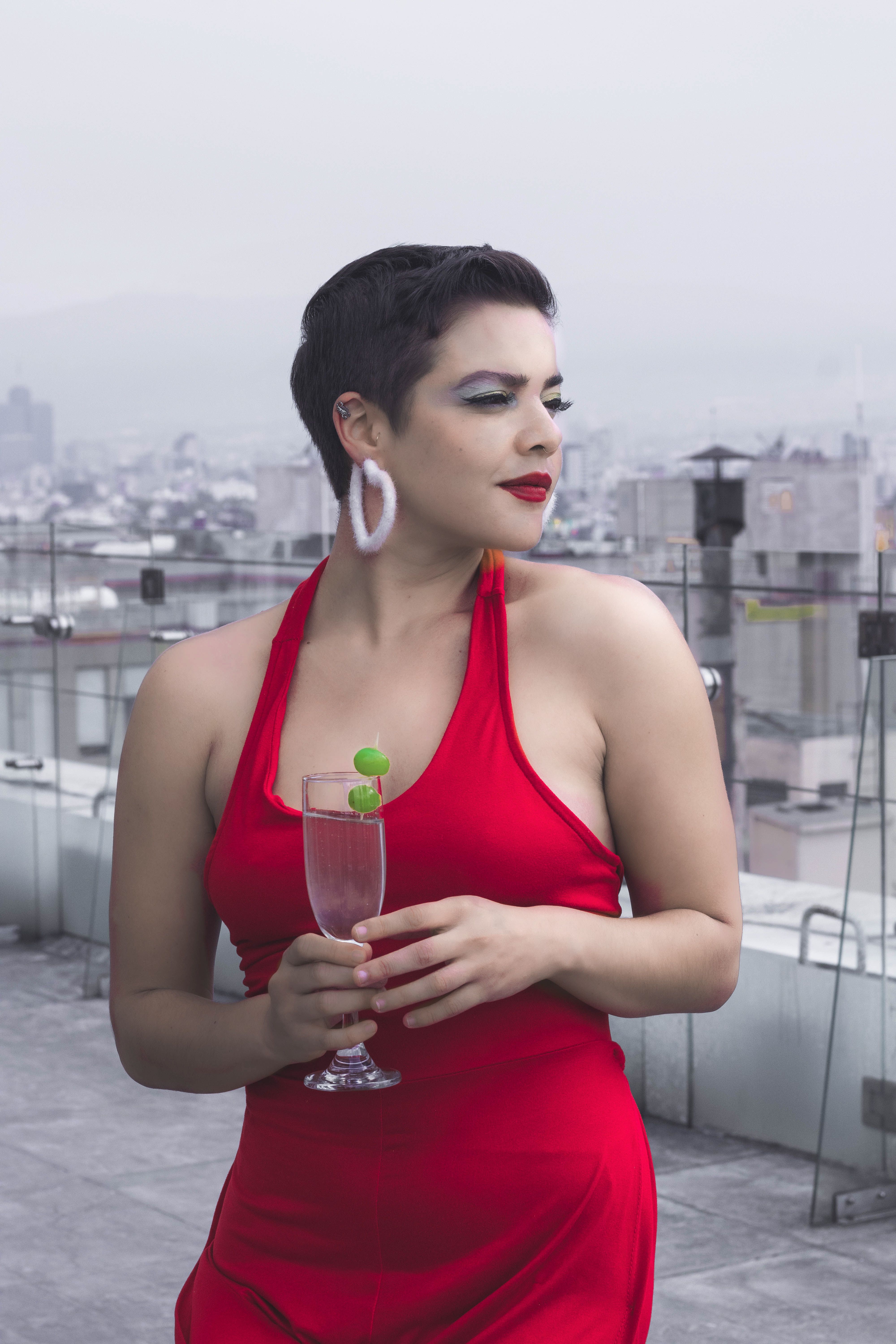 Elegant woman in a red dress enjoys a drink on a city rooftop, embodying luxury and fashion.