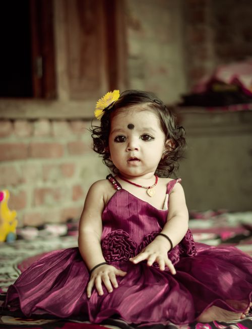 Free Charming baby girl in a purple dress sits indoors with a flower in her hair, showcasing innocence and beauty. Stock Photo