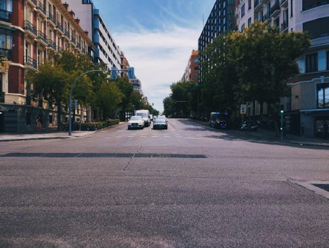 Wide urban street intersection bordered by tall buildings and trees under a clear sky.