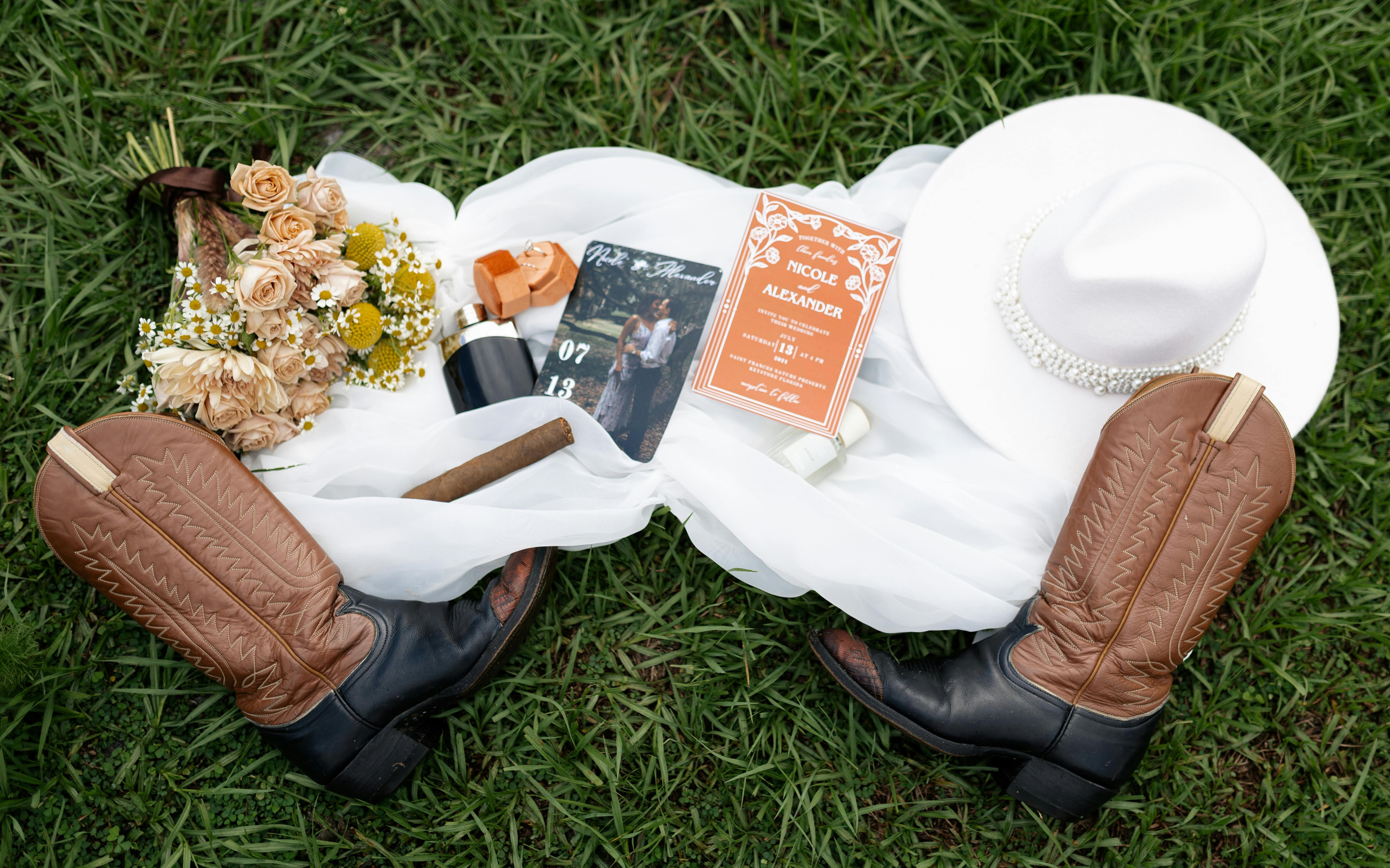 Country Wedding Music: Songs, DJs & Planning Tips - Flat lay of wedding items including cowboy boots, a white hat, and floral bouquet on grass.