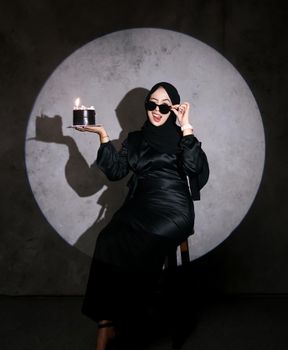 Woman in sunglasses and hijab holding a small cake with lit candles under a spotlight.