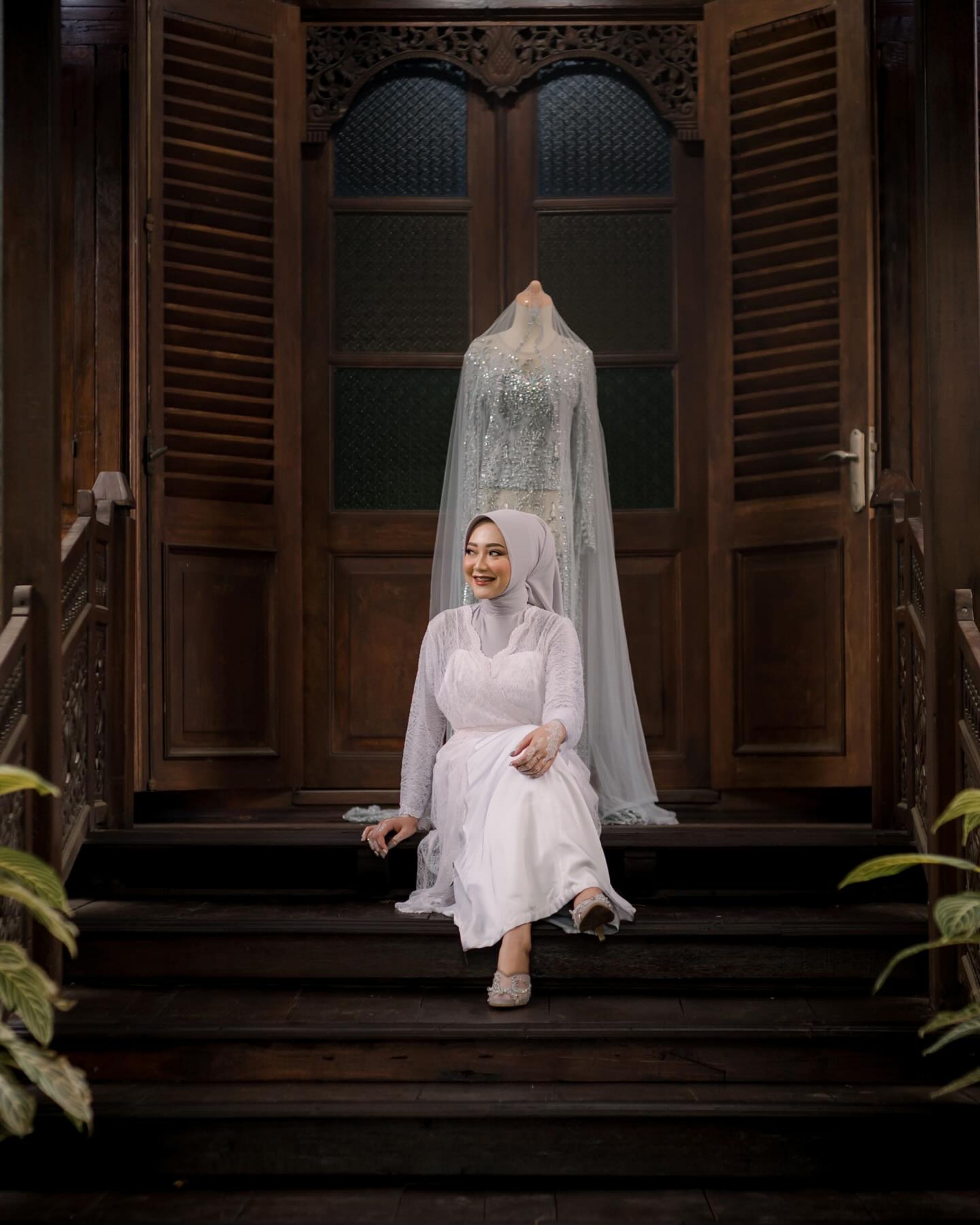 An elegant bride in a traditional white gown sits gracefully in front of ornate wooden doors, embodying serenity and style.