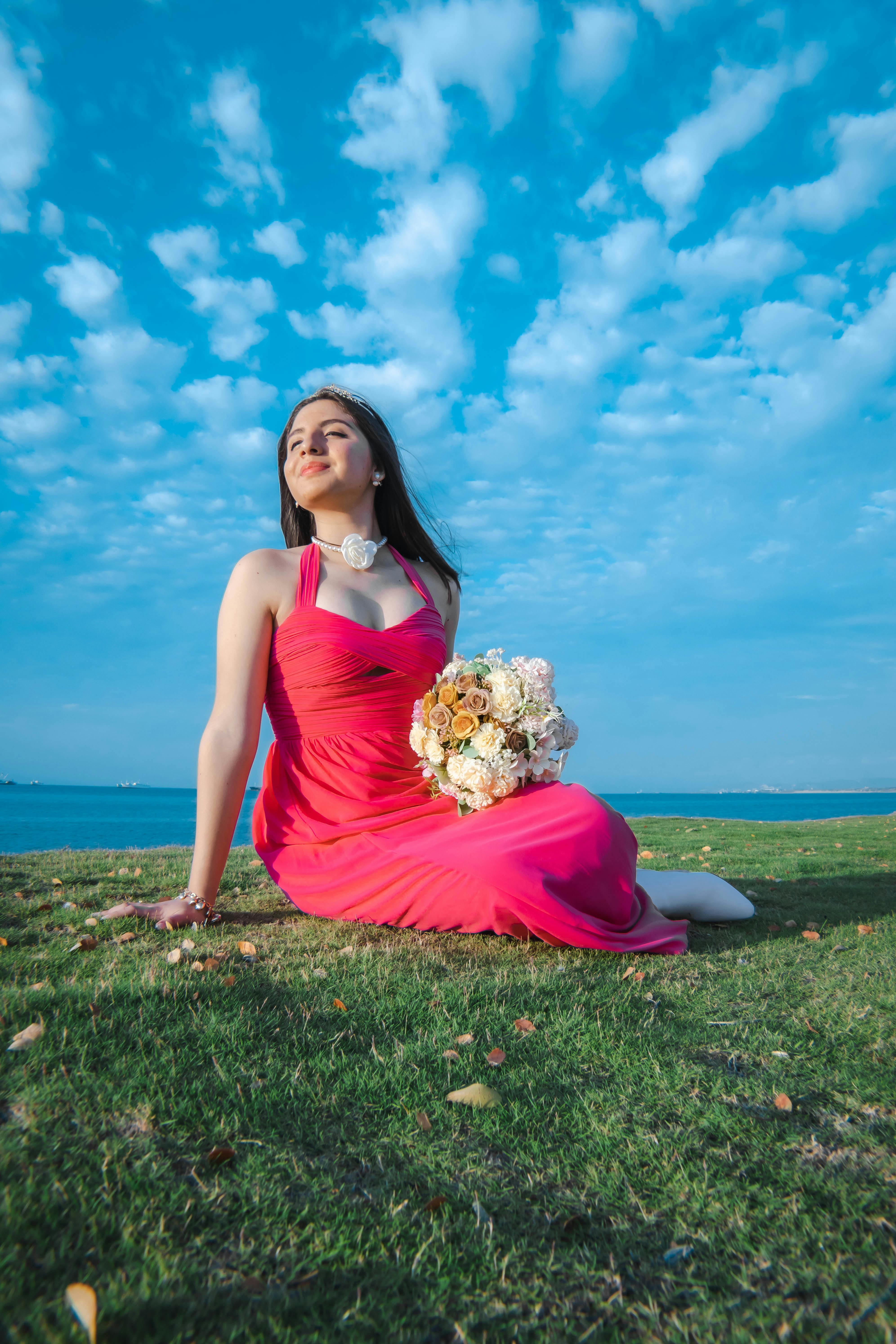 A joyful woman in a vibrant pink dress sits on grass, holding a bouquet against a clear blue sky.