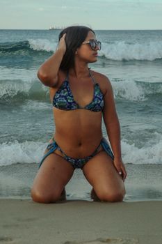 Woman in a bikini kneeling on a sandy beach with ocean waves in the background, enjoying a summer day.