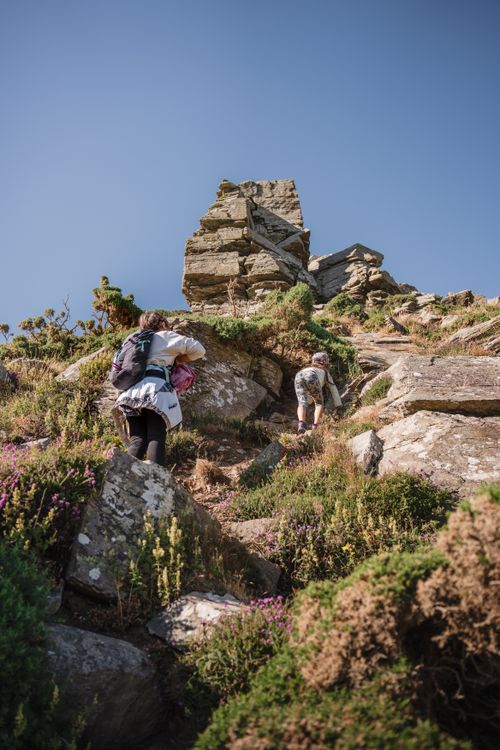Free Two hikers climbing a rugged cliffside in daylight, surrounded by lush vegetation, under a clear summer sky. Stock Photo