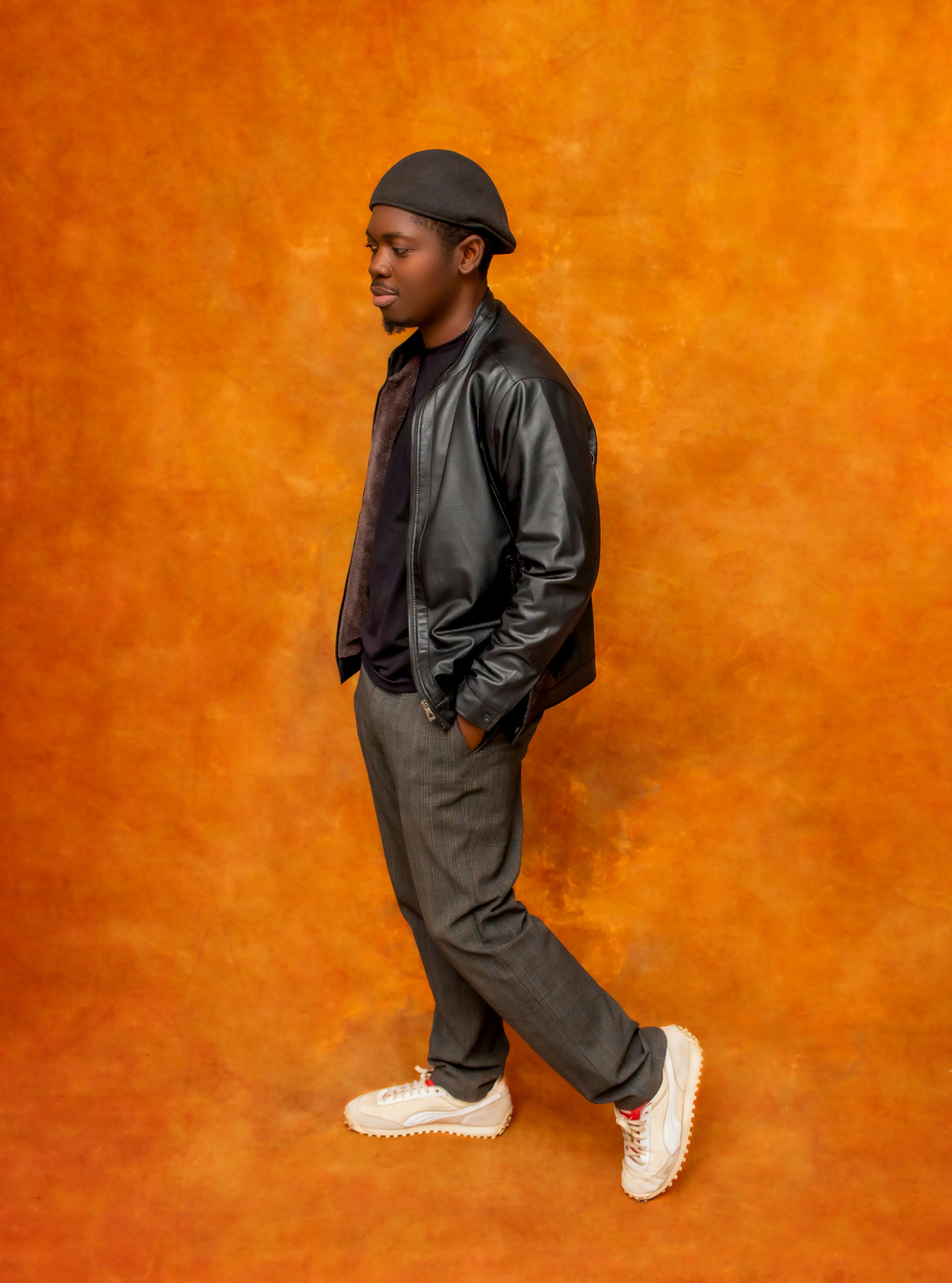 Fashionable man poses confidently in a leather jacket on a vibrant orange background.