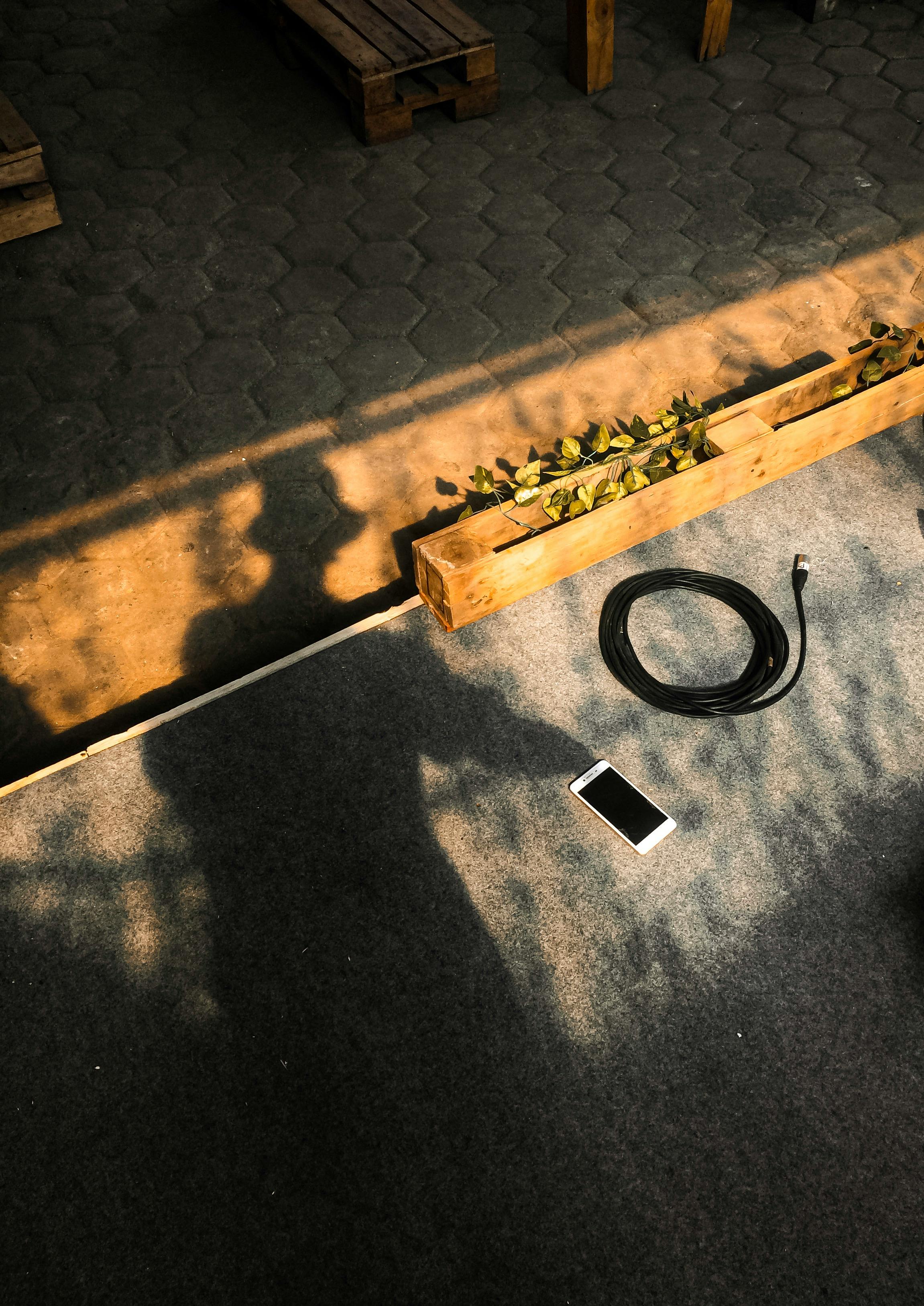 A smartphone and cable on a sunlit pavement with shadows of trees.