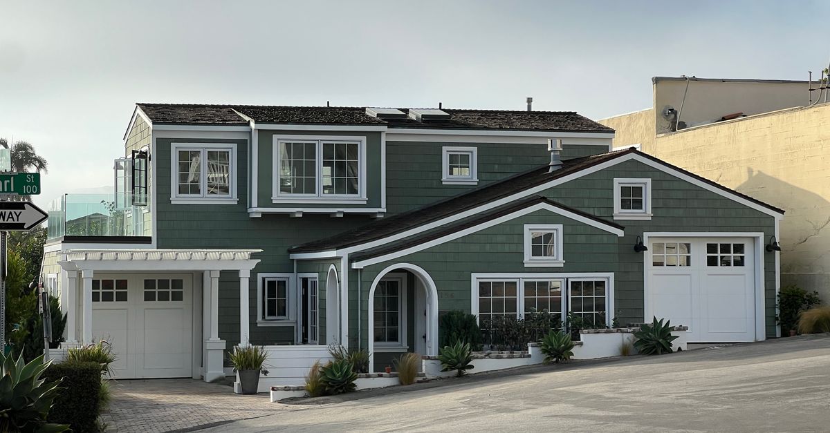 A stylish green two-story suburban house with white trim and garages on a sunny street corner.
