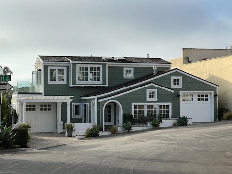 A stylish green two-story suburban house with white trim and garages on a sunny street corner.