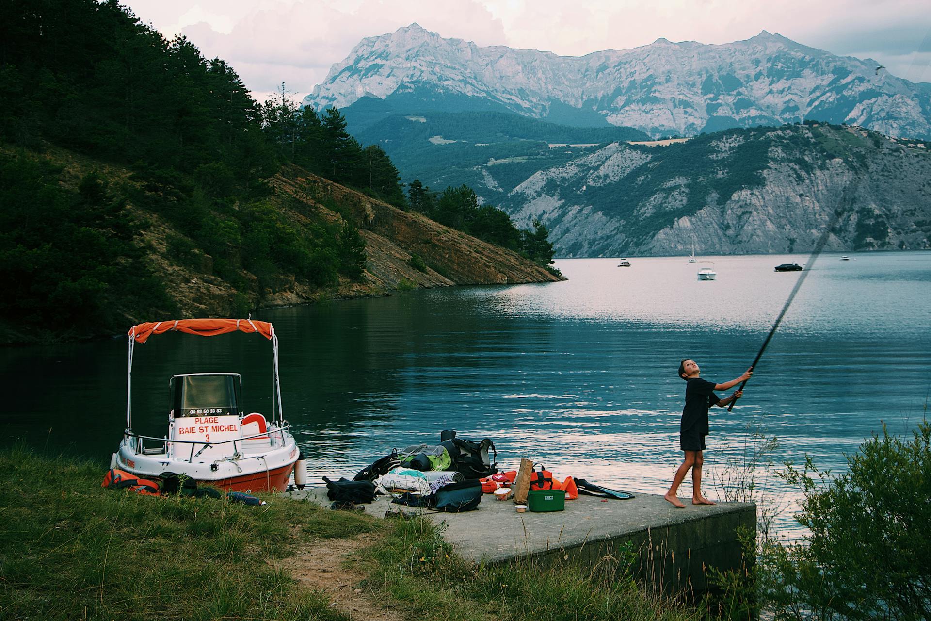 Young boy fishing at a scenic mountain lake with a boat nearby in summer.