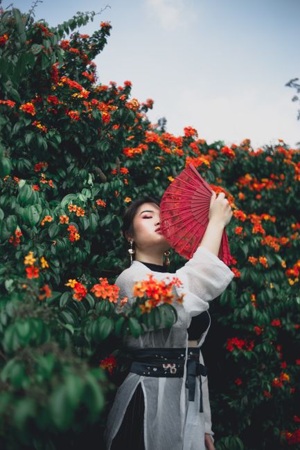 Stylish woman posing with red fan amidst colorful garden blooms, blending elegance with nature.