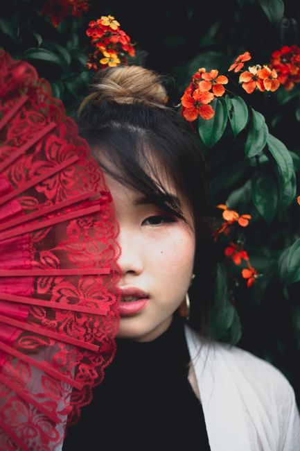 Close-up portrait of a woman partially covered with a red lace fan, surrounded by vibrant flowers.