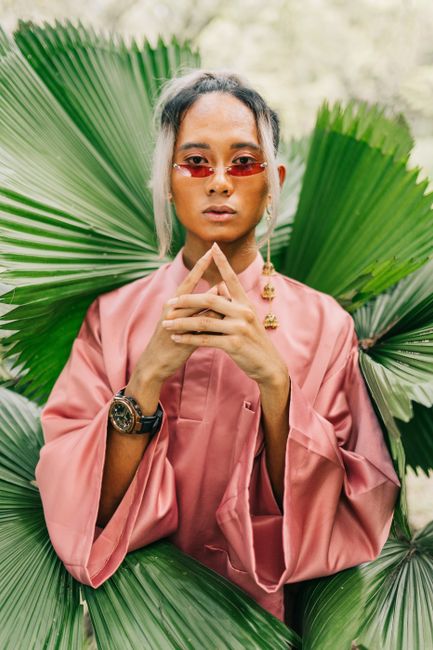 Stylish model posing with red sunglasses and pink outfit surrounded by palm leaves.