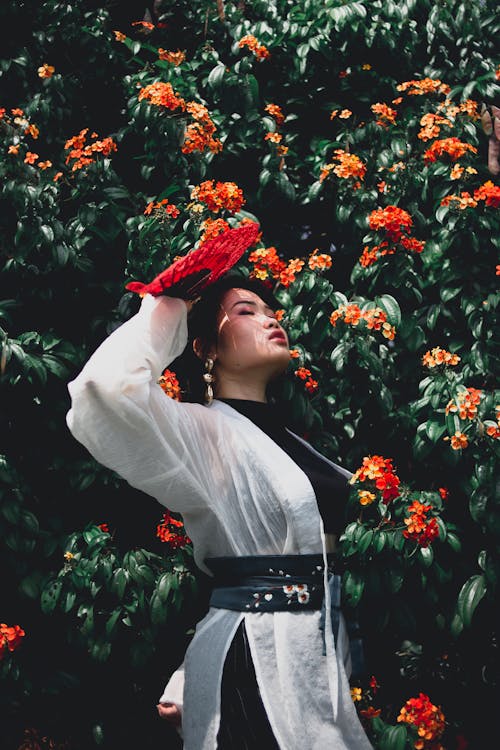 Side view of Asian woman in black and white outfit with fan above head standing against flowering plant