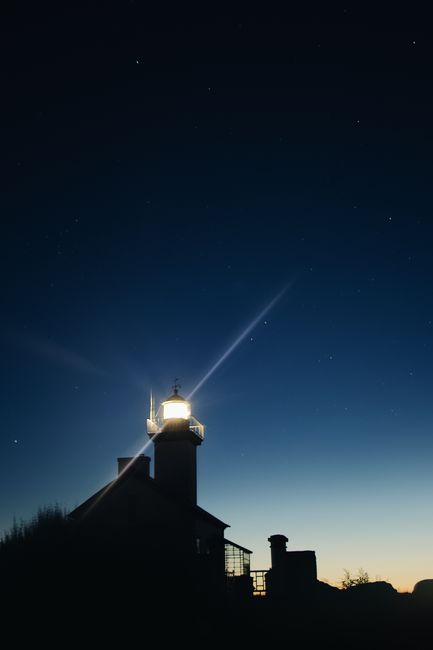 A silhouetted lighthouse illuminated at twilight beneath a starry sky, offering a serene coastal night view.