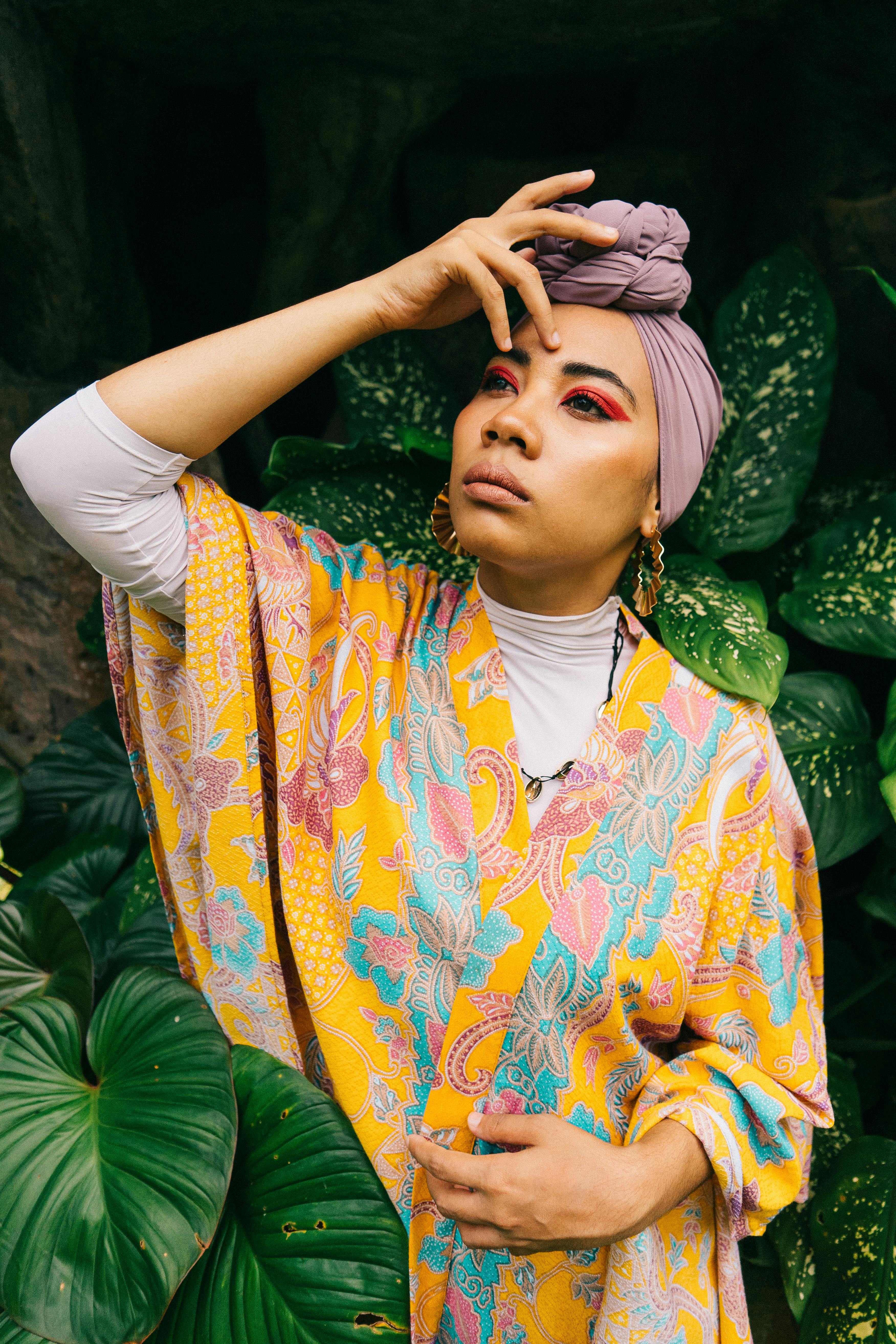 Fashionable woman in vibrant attire poses with expressive makeup and headscarf amidst lush green leaves.
