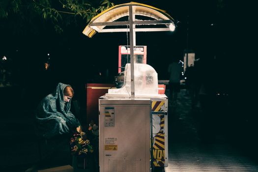 A woman wrapped in a blanket sits silently next to a street vendor cart at night.